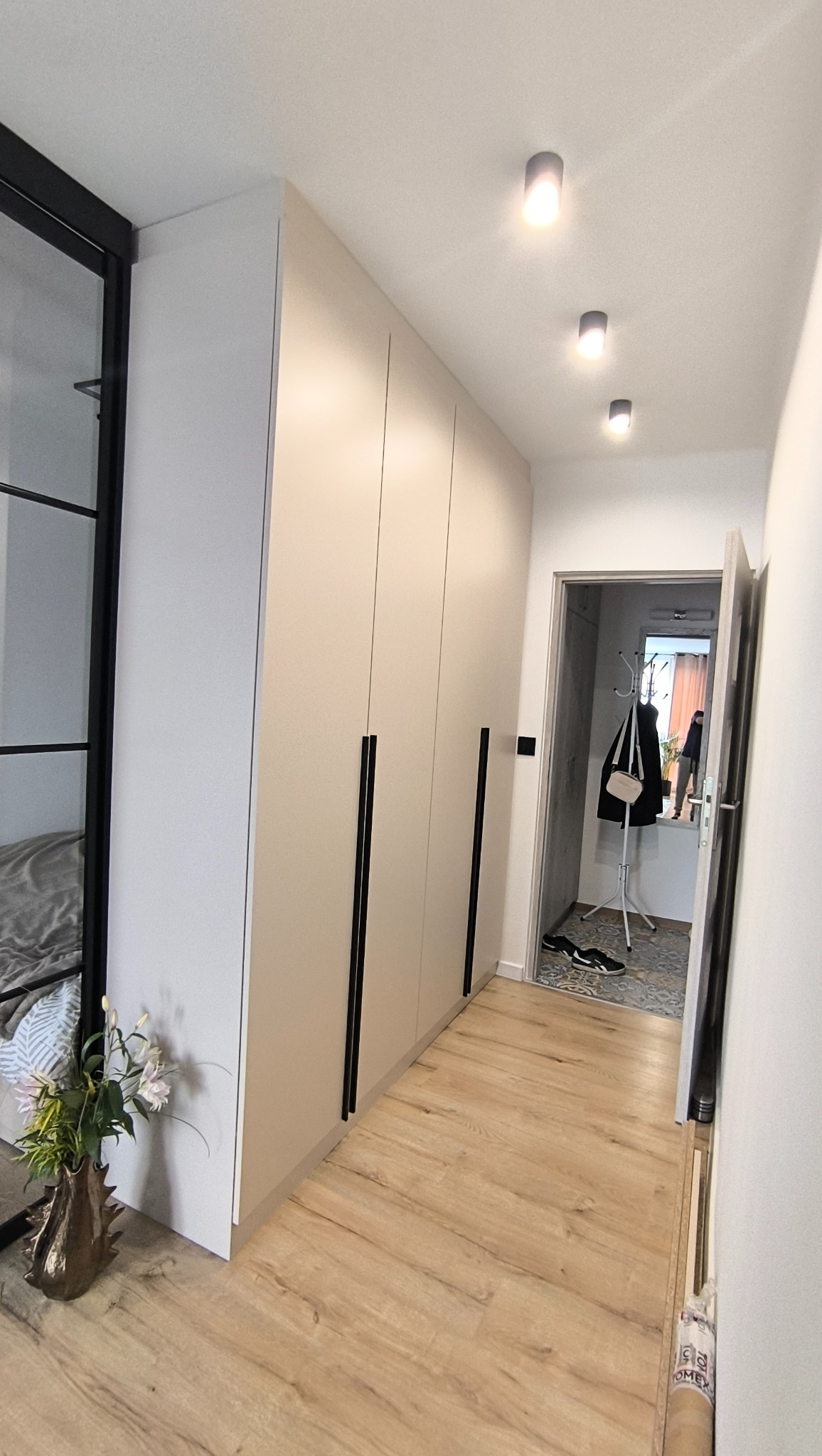 Beżowa szafa wnękowa z czarnymi uchwytami, widok na przedpokój z drewnianą podłogą i szklane drzwi. Minimalistyczny design, oświetlenie sufitowe punktowe.