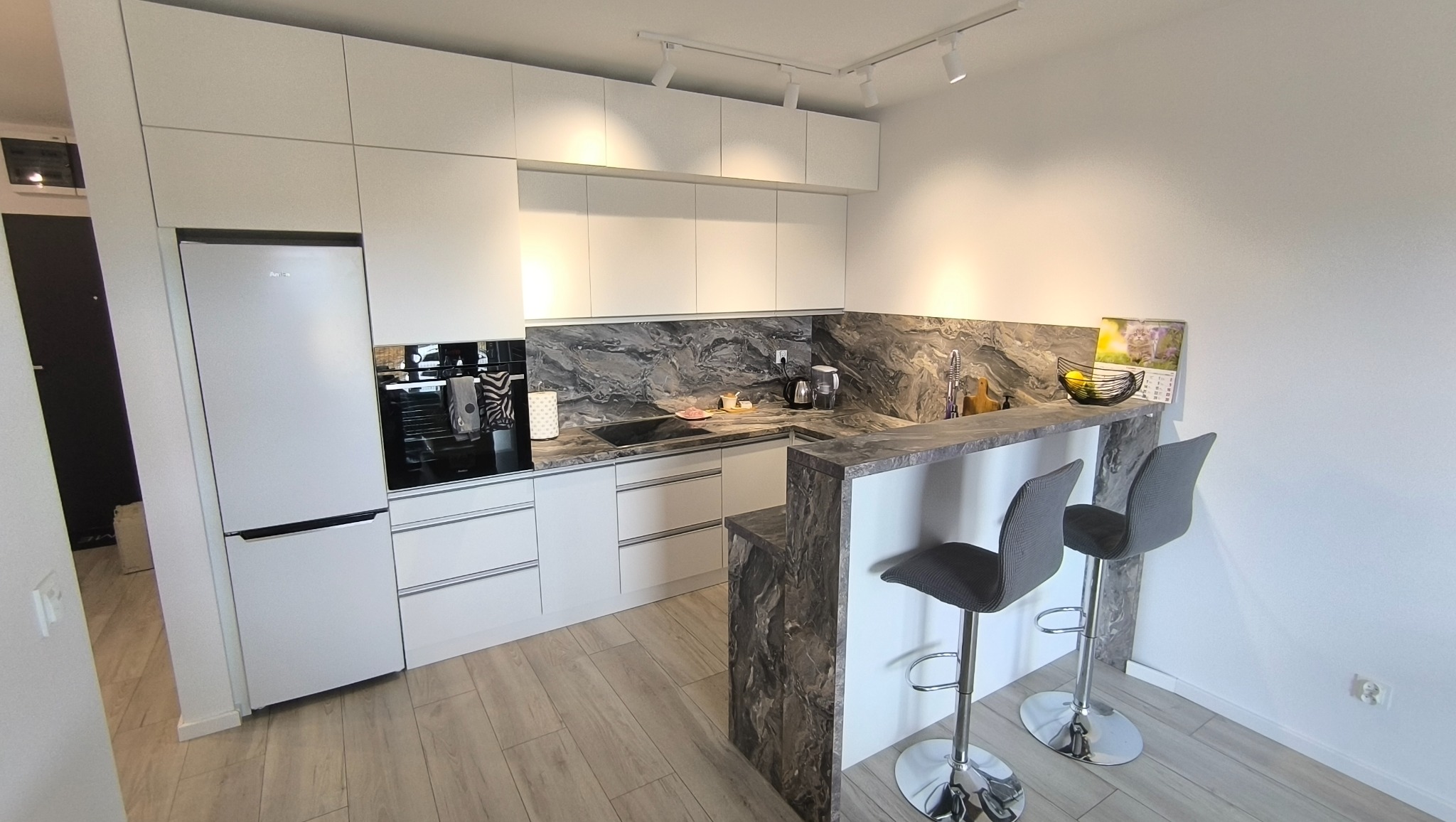 Nowoczesna kuchnia na wymiar z białymi frontami, blatem imitującym marmur i barkiem z dwoma hokerami. Oświetlenie punktowe.