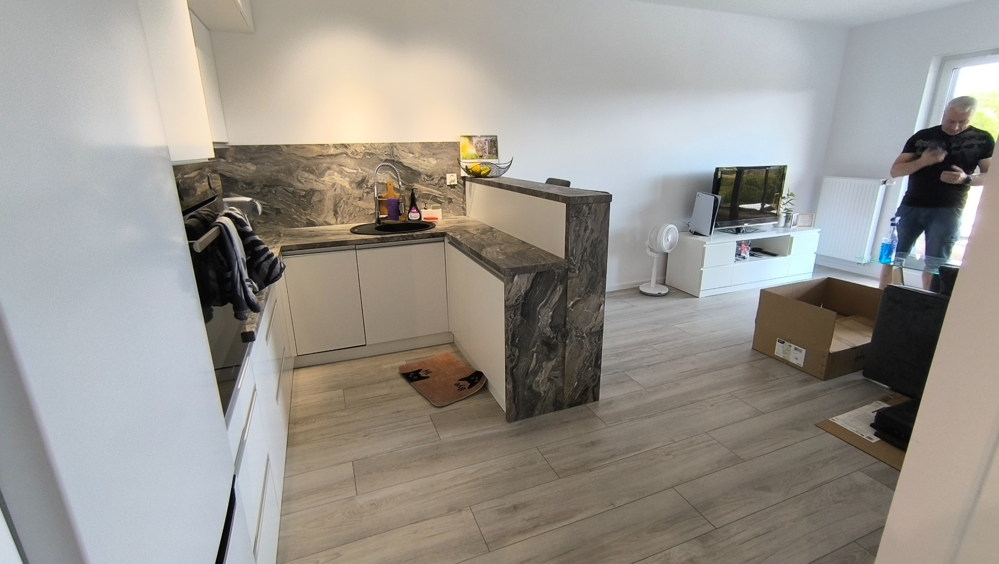 Nowoczesna kuchnia z białymi szafkami i blatem imitującym marmur, widoczna z perspektywy wejścia do pokoju. Uwagę przykuwa wykończenie półwyspu marmurkowym wzorem.