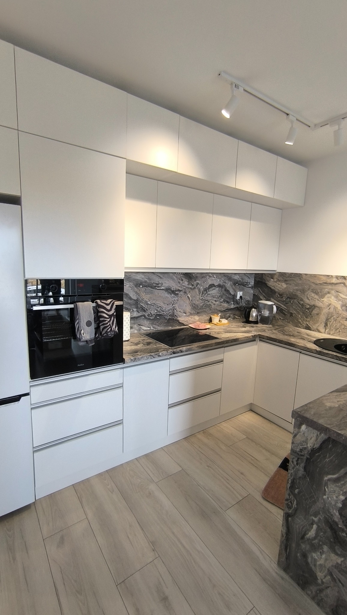 Nowoczesna, biała kuchnia z blatem imitującym marmur, oświetleniem punktowym i drewnianą podłogą. Widoczny piekarnik z ręcznikami i elementy wyposażenia.