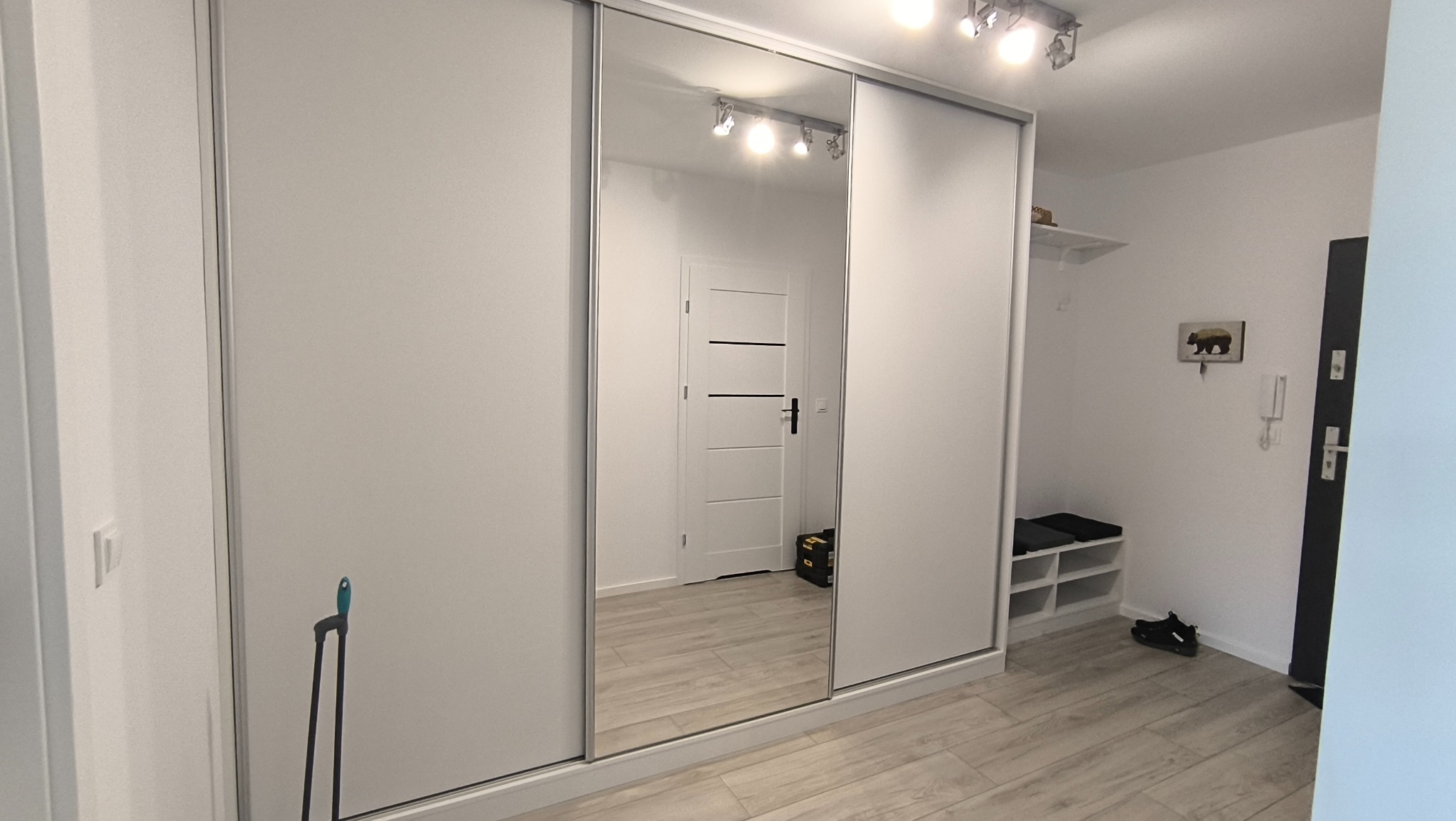 Biała szafa wnękowa z lustrzanymi drzwiami przesuwnymi, odbijająca wnętrze przedpokoju z białą podłogą i drzwiami. Minimalistyczny design.