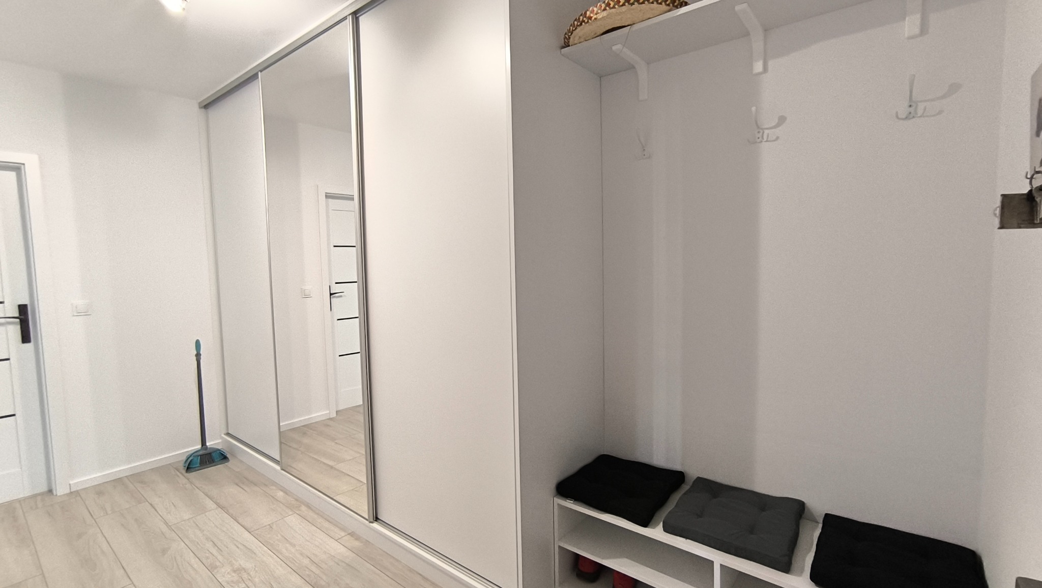 Biała szafa wnękowa z lustrzanymi drzwiami przesuwnymi w przedpokoju. Obok wieszak z siedziskiem i półkami na buty. Minimalistyczny design, jasne wnętrze.