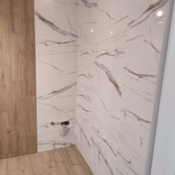 Marek Podstawka - Wykończona łazienka z drewnopodobną podłogą i ścianami w płytkach imitujących marmur z brązowymi żyłami. Widoczne przyłącza wodne.