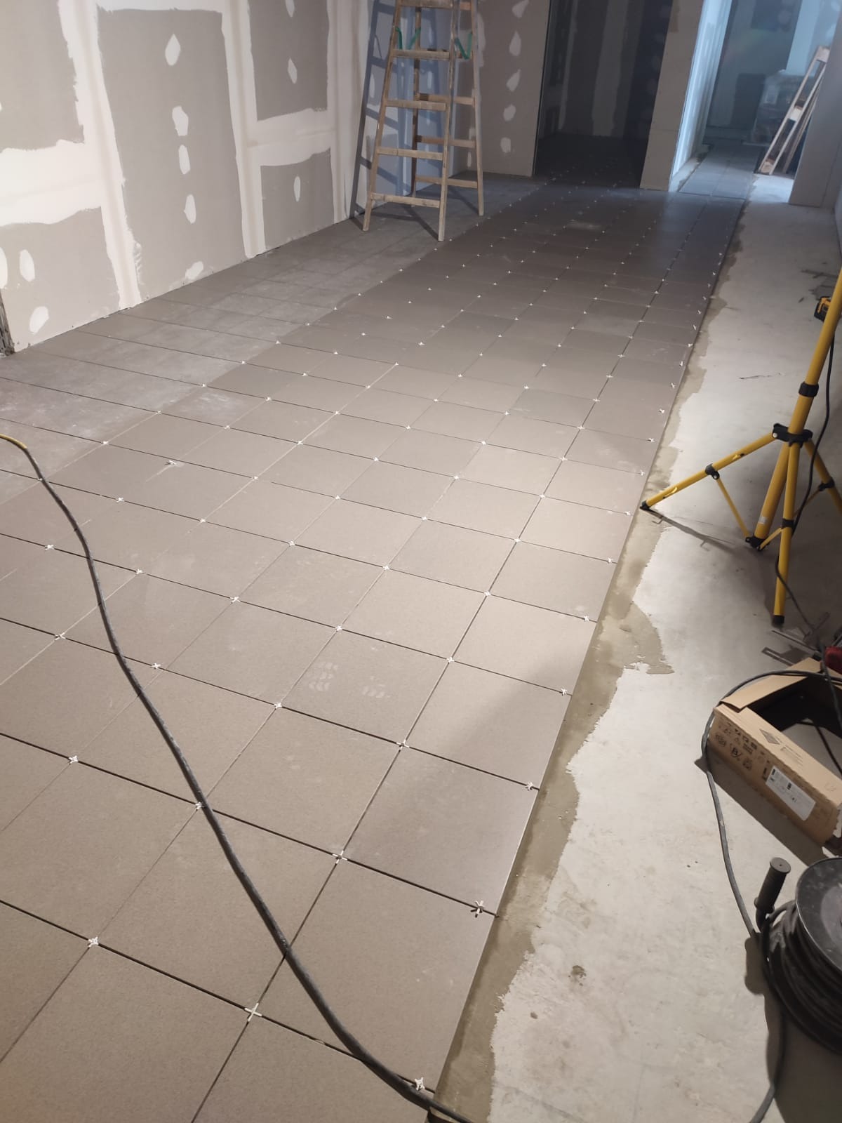 Układanie beżowych płytek podłogowych z krzyżykami dystansowymi. W tle drabina i ściany przygotowane do malowania.