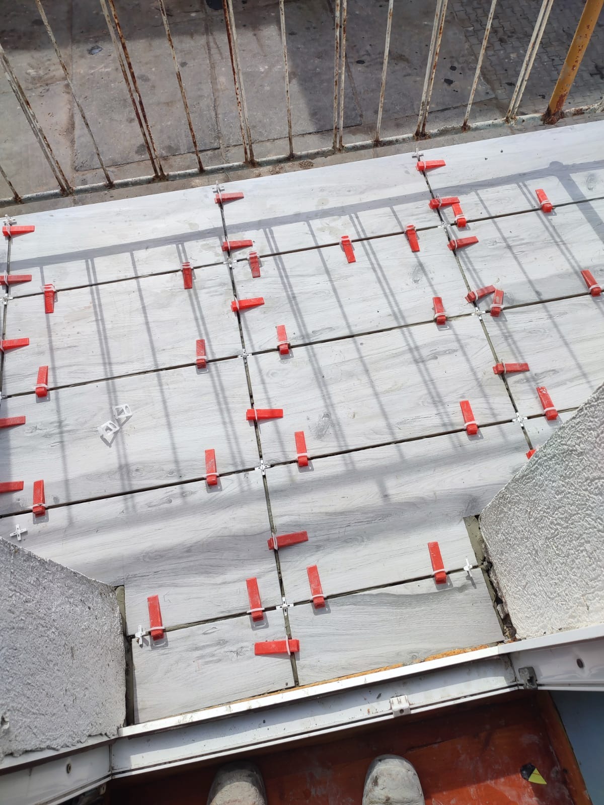 Jasnoszare płytki imitujące drewno na balkonie, wyrównane czerwonymi klipsami i krzyżykami dystansowymi. Widok z góry, fragment balustrady i butów.