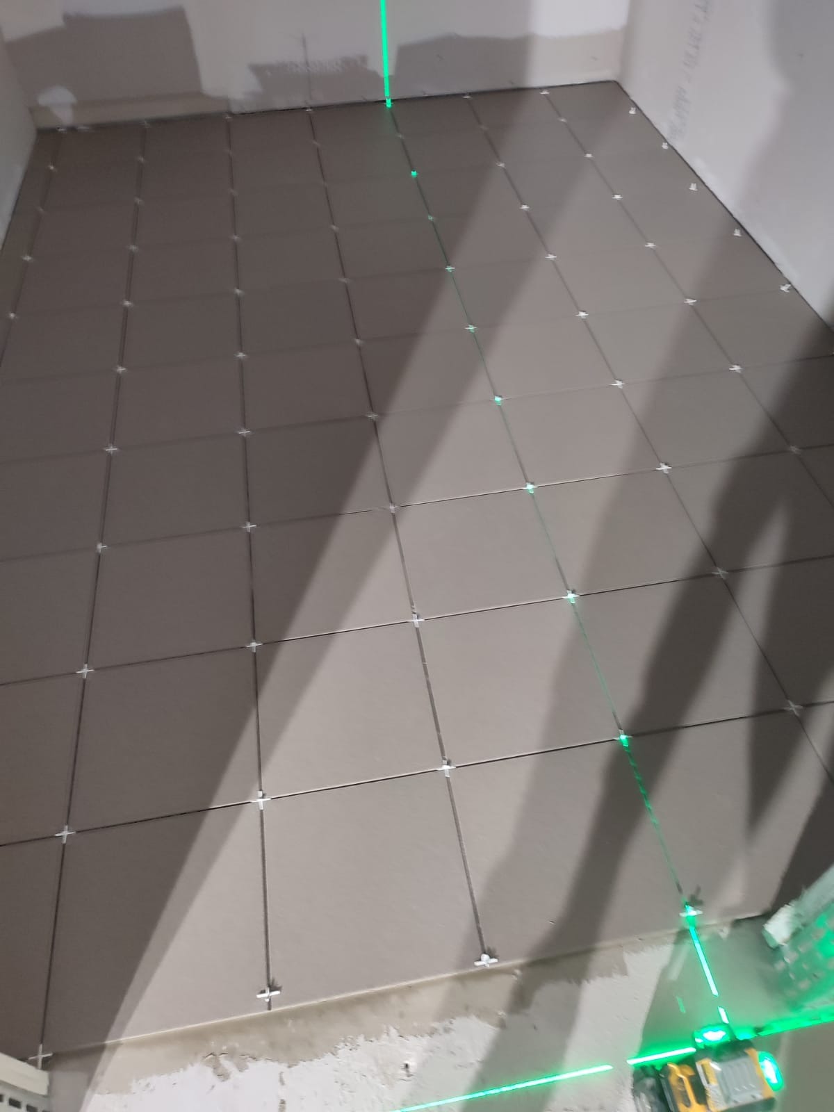 Układanie szarych płytek podłogowych z użyciem poziomicy laserowej i krzyżyków dystansowych. Widoczne fugi i fragment ściany w surowym stanie.