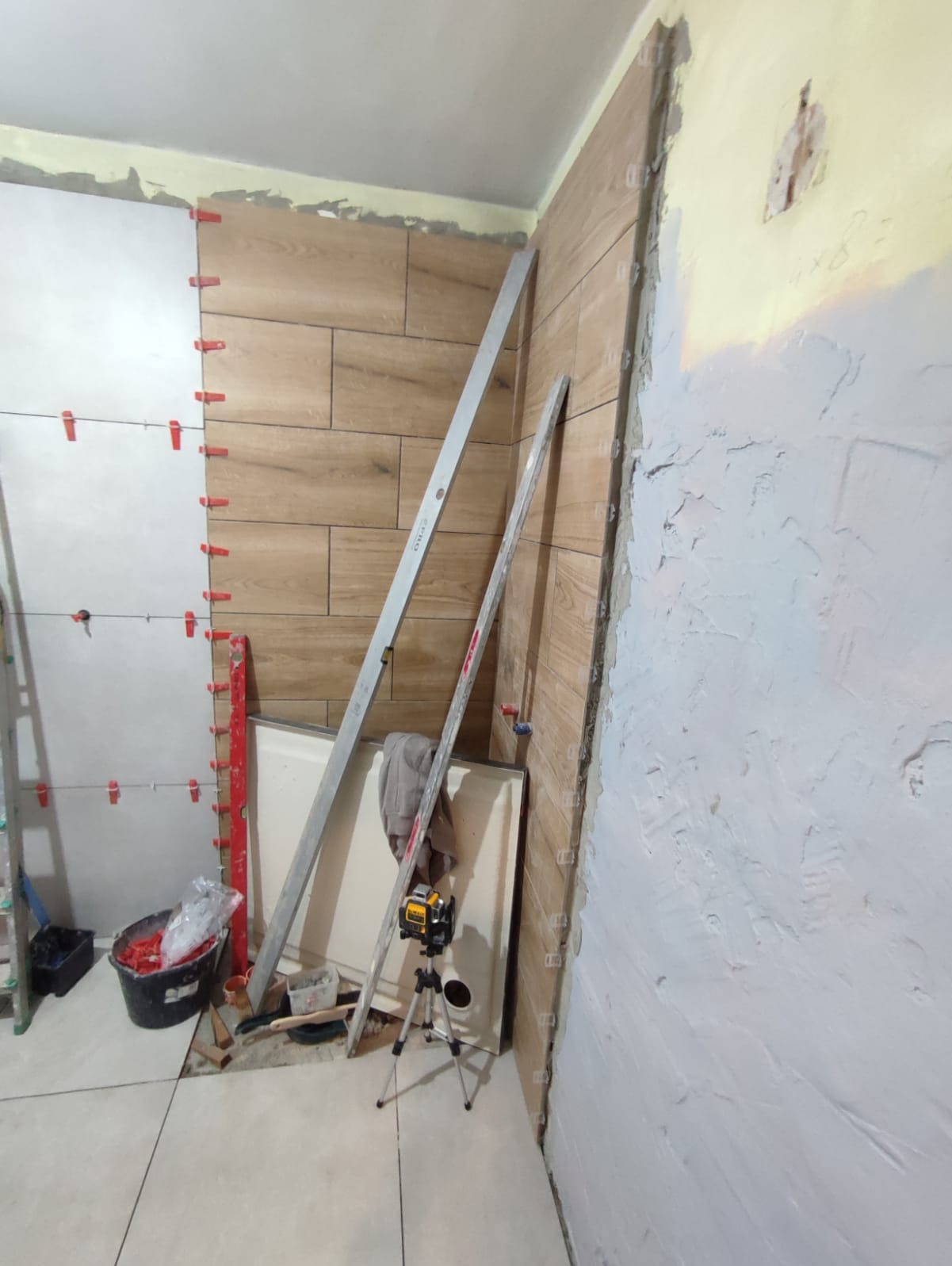 Montaż płytek imitujących drewno na ścianie w łazience, z użyciem poziomicy laserowej i klipsów poziomujących. Częściowo położone płytki, surowa ściana w tle.