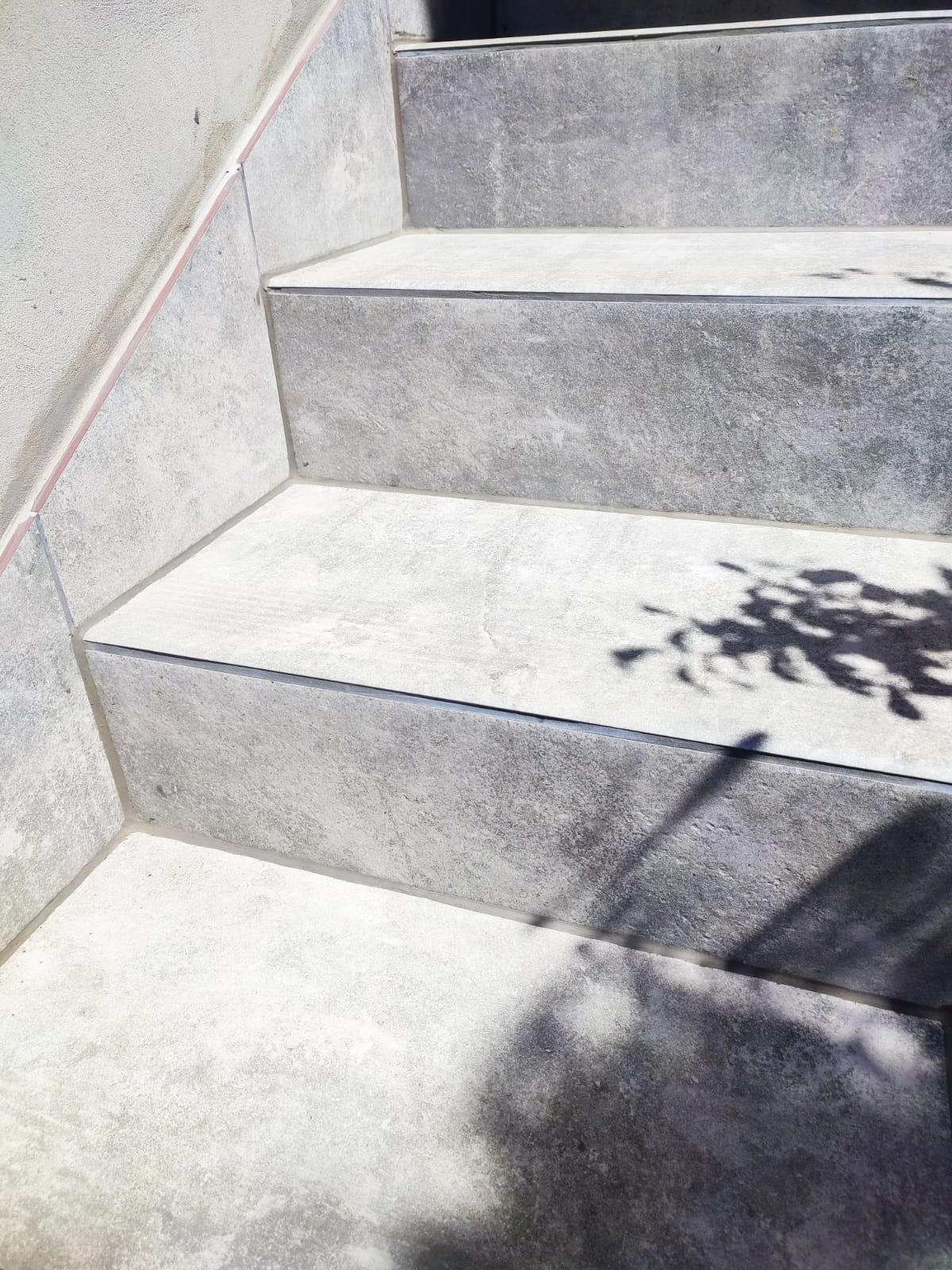 Wykończone schody zewnętrzne z szarych, matowych płytek, zabezpieczone listwami. Na płytkach cień rzucany przez liście. Estetyczne połączenie materiałów.