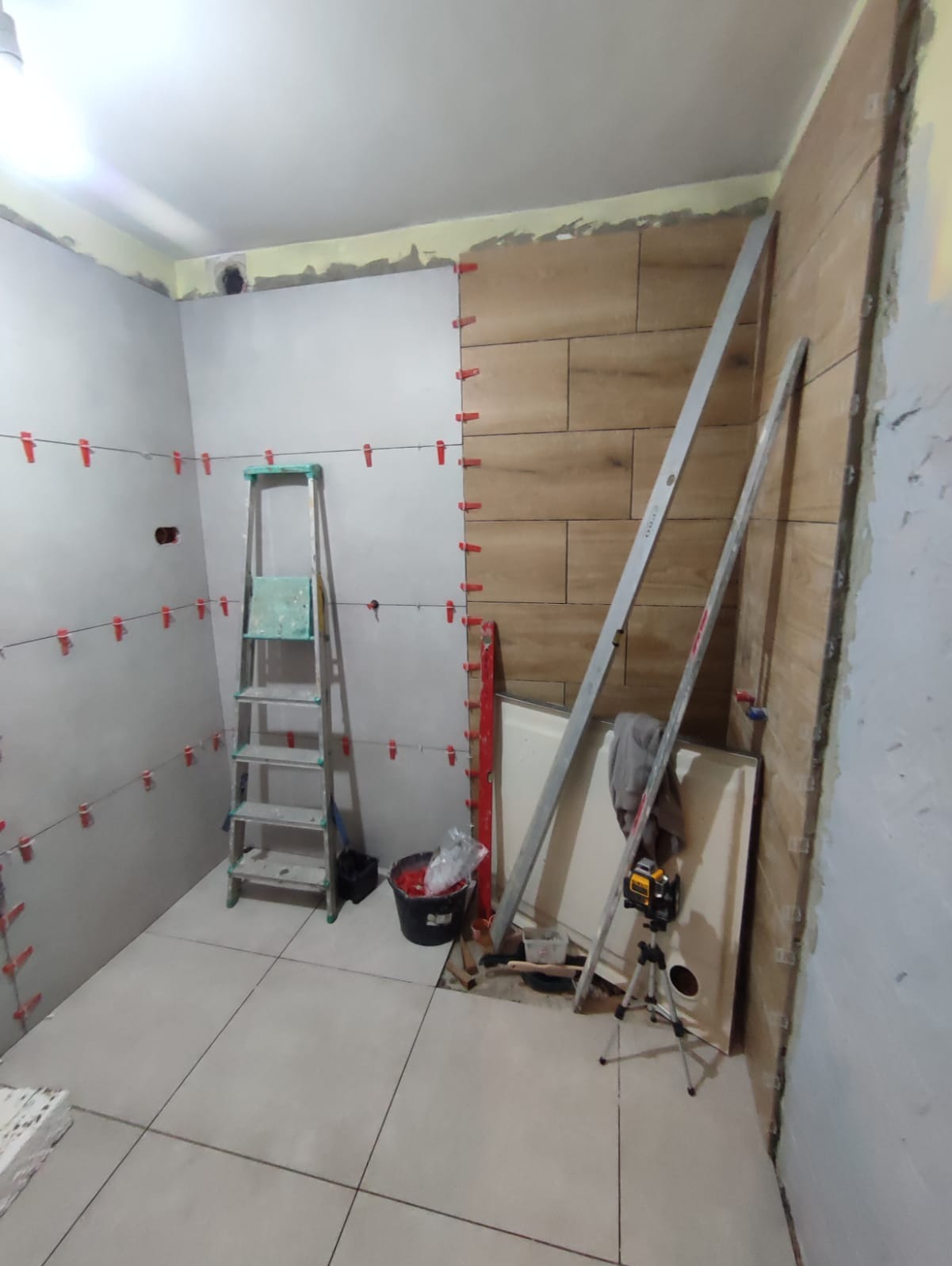 Układanie płytek drewnopodobnych na ścianie łazienki. Widoczne poziomice, drabina i system poziomowania płytek. Częściowo ułożona podłoga z jasnych płytek.