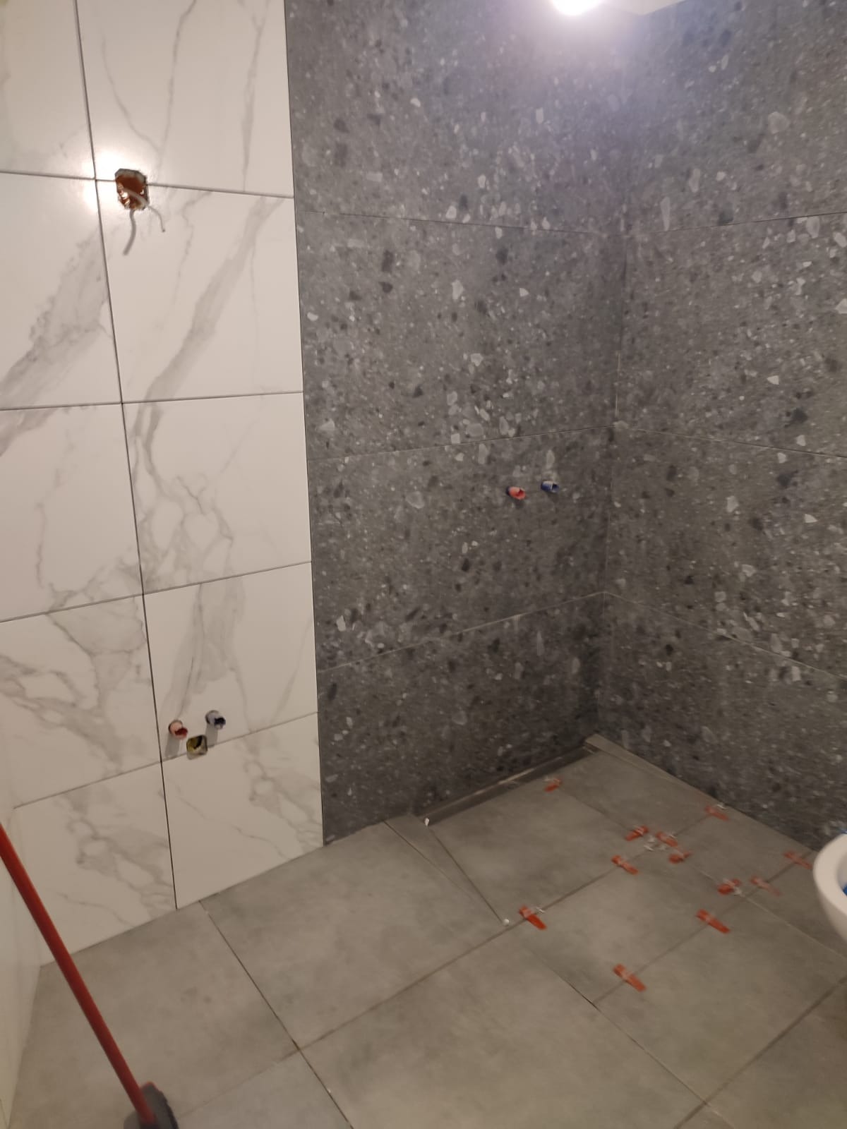 Łazienka w trakcie wykańczania: białe płytki imitujące marmur zestawione z szarymi płytkami o nieregularnej fakturze. Widoczne krzyżyki dystansowe.