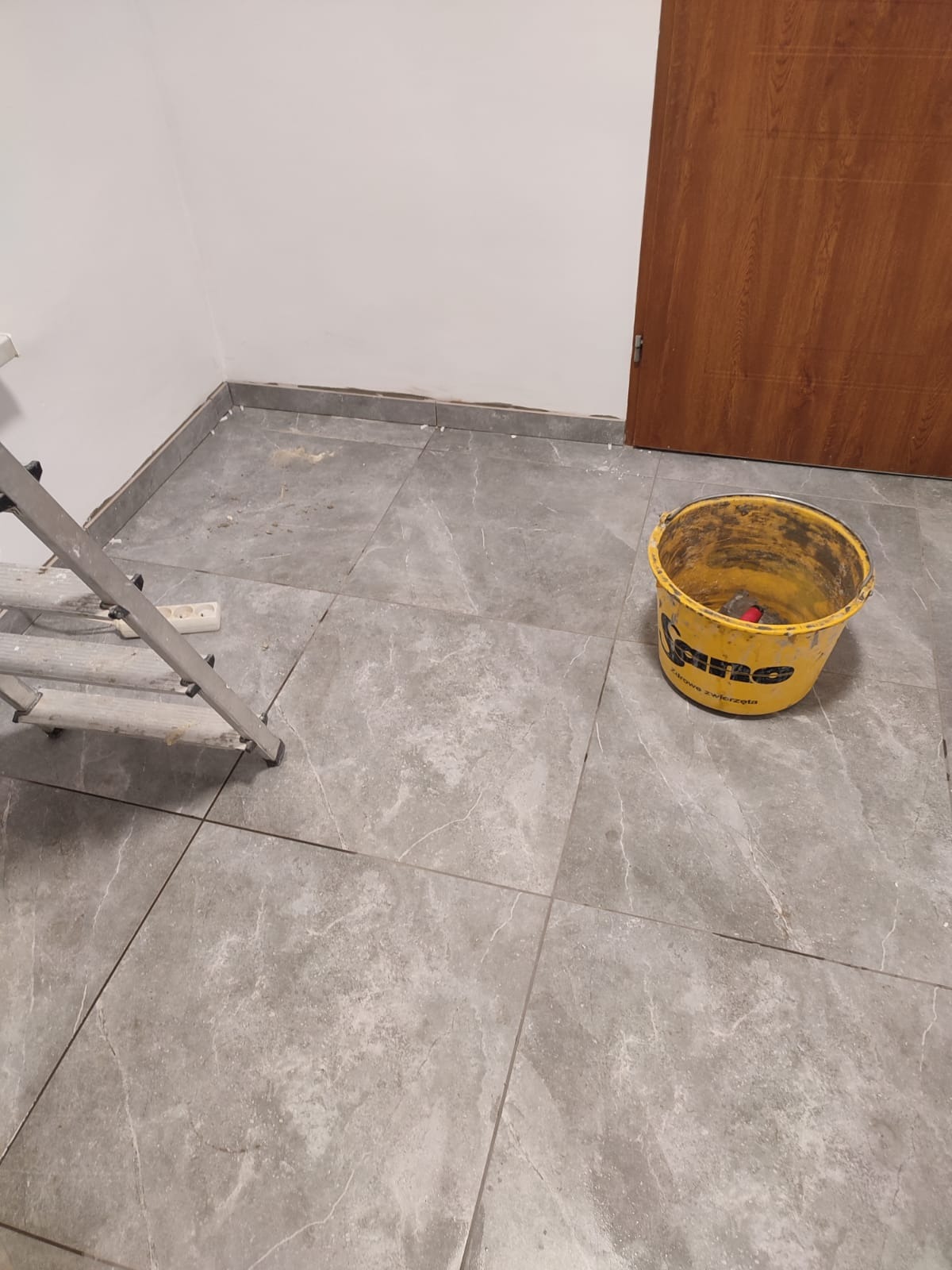 Ułożone szare płytki podłogowe w pomieszczeniu, widoczne wiadro z resztkami zaprawy i fragment drabiny. Praca w toku.
