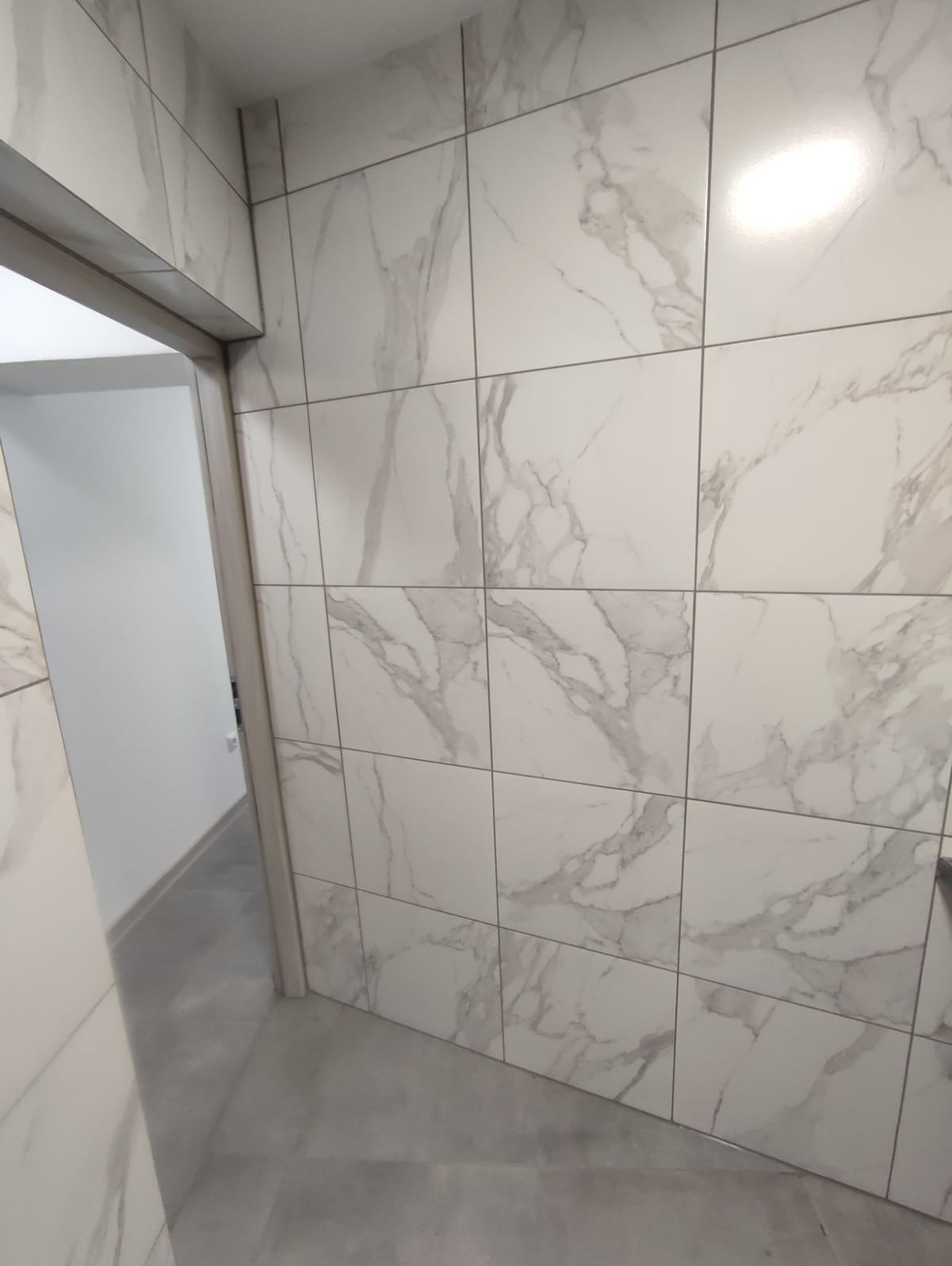 Ściana wyłożona białymi, kwadratowymi płytkami z szarym marmurkowym wzorem, widoczny fragment futryny drzwi i szara podłoga. Minimalistyczne wykończenie wnętrza.