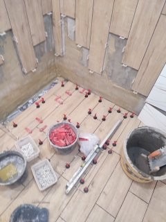 Układanie płytek imitujących drewno w rogu pomieszczenia. Widoczne narzędzia glazurnicze i system poziomowania płytek. Częściowo przygotowana ściana.