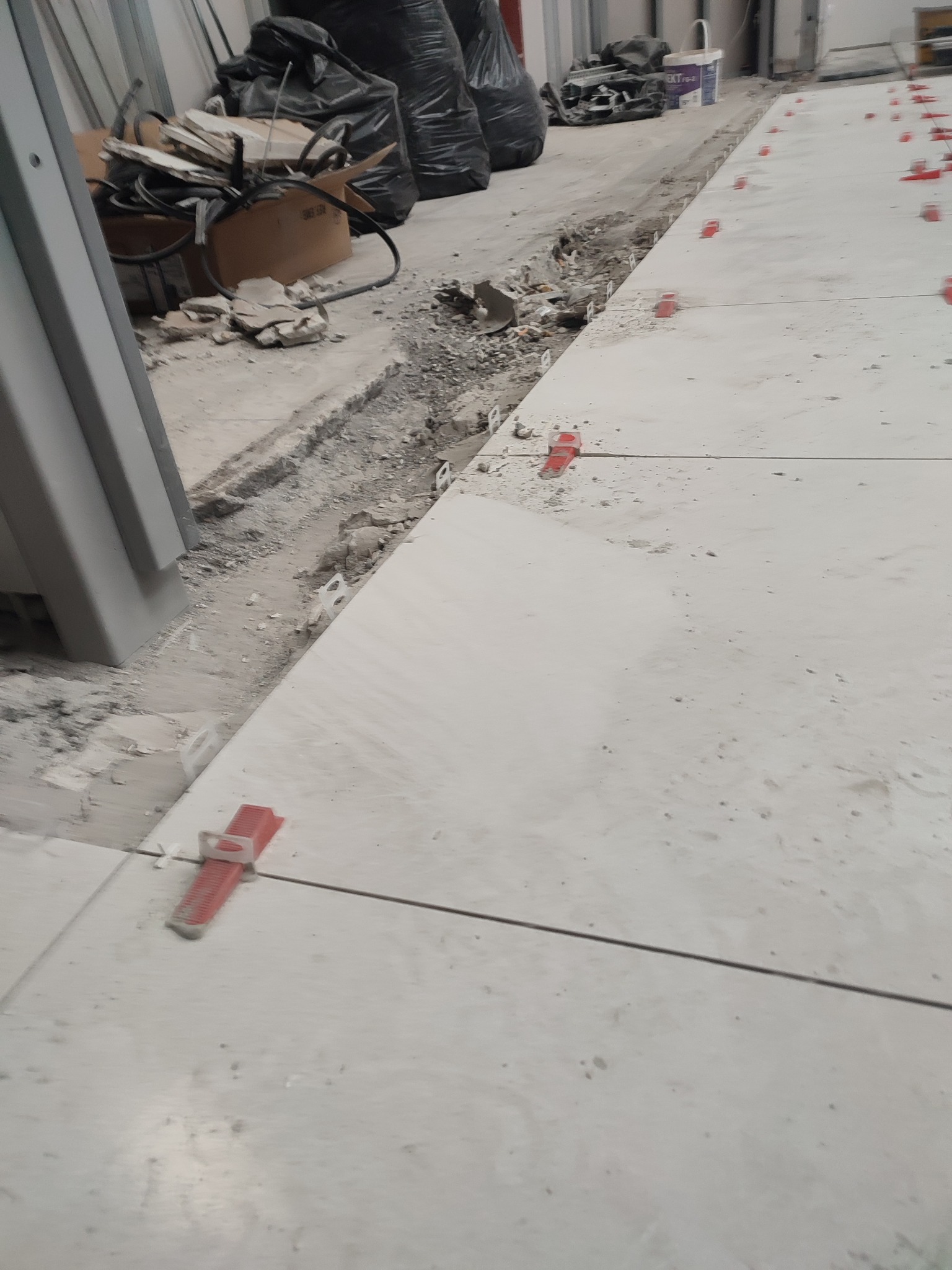 Układanie jasnych płytek podłogowych z użyciem czerwonych klipsów poziomujących. Widoczne ślady prac remontowych i gruz.