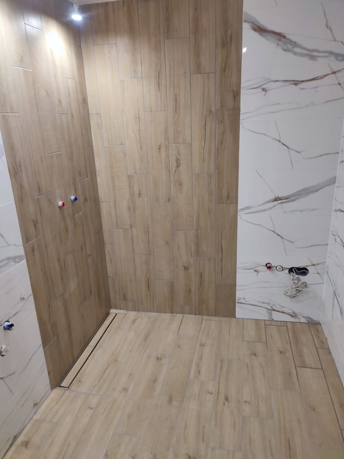 Wykończona kabina prysznicowa z płytkami imitującymi drewno i marmur. Widoczne odpływ liniowy i wystające przyłącza wodne.