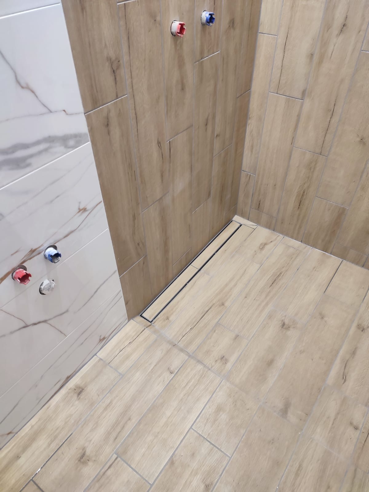 Ukończona kabina prysznicowa z odpływem liniowym i płytkami imitującymi drewno na ścianach. Widoczne przyłącza wody. Jasne, naturalne światło.