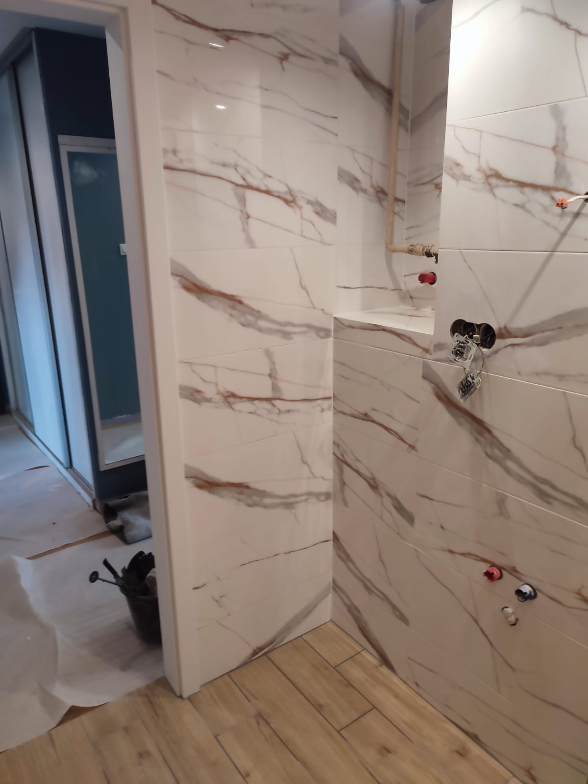 Ściana łazienki wyłożona białymi płytkami imitującymi marmur, z widoczną instalacją wodną i elektryczną. Na podłodze panele imitujące drewno. Częściowo widoczne narzędzia.