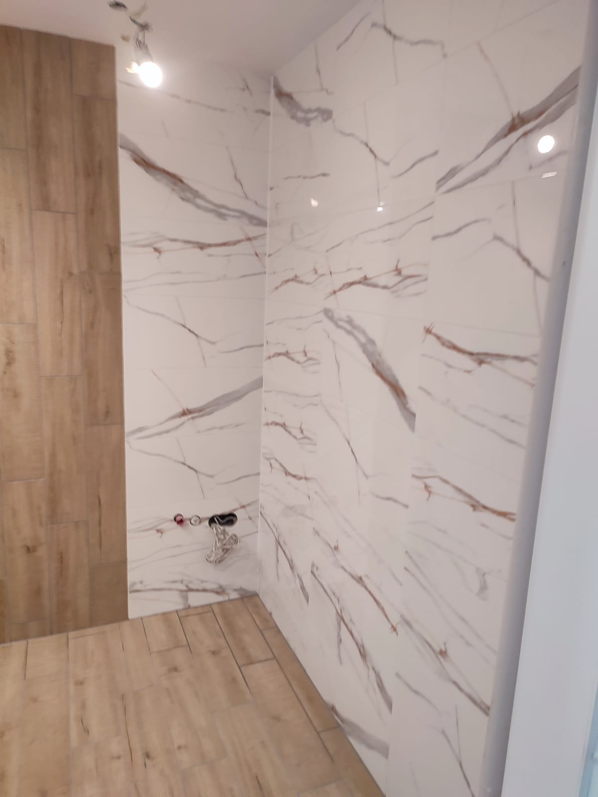 Wykończona łazienka z drewnopodobną podłogą i ścianami w płytkach imitujących marmur z brązowymi żyłami. Widoczne przyłącza wodne.