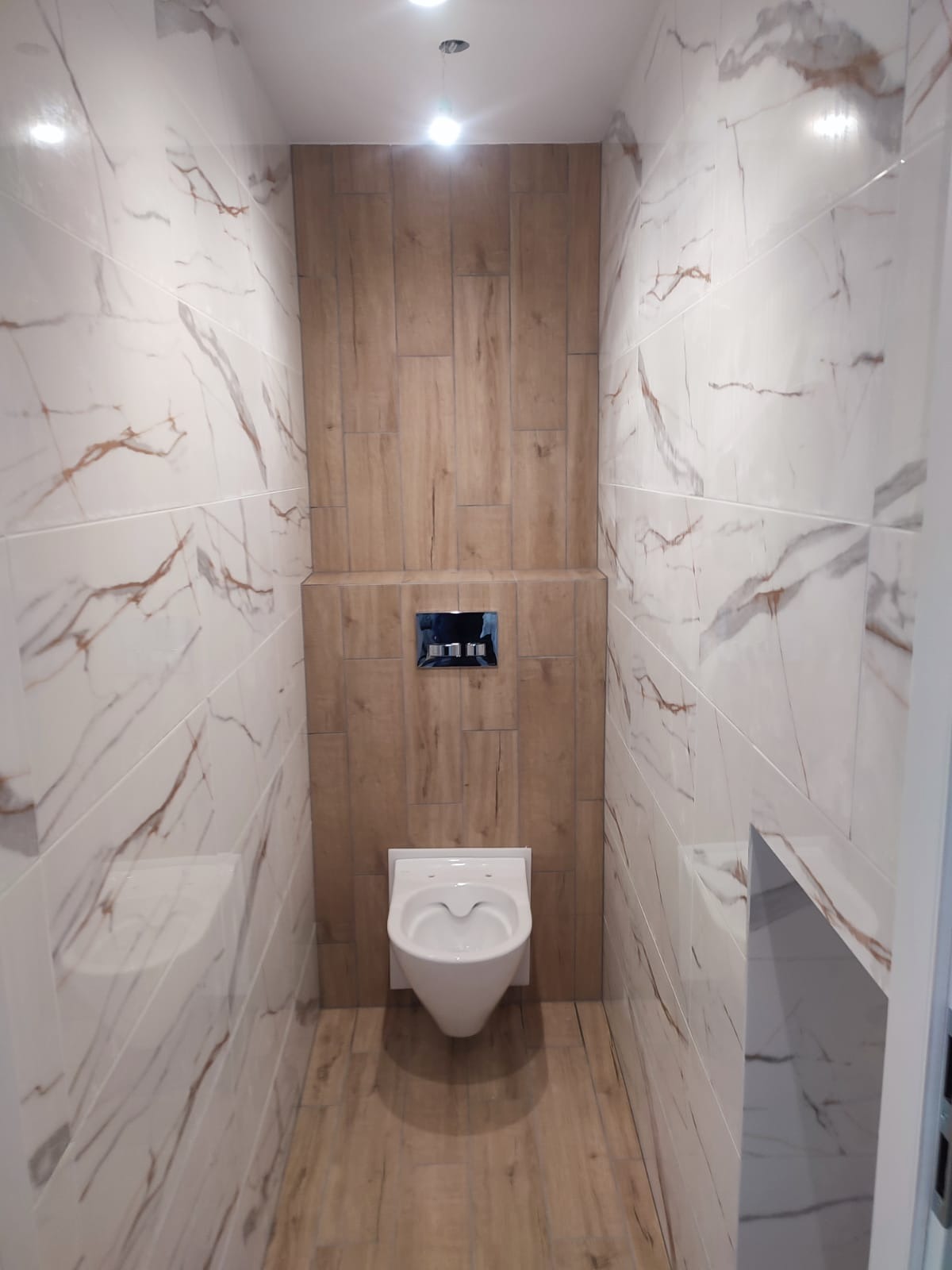 Nowoczesna toaleta w beżowo-białej kolorystyce, z płytkami imitującymi marmur i drewno. Podwieszana miska WC i minimalistyczny przycisk spłukujący.