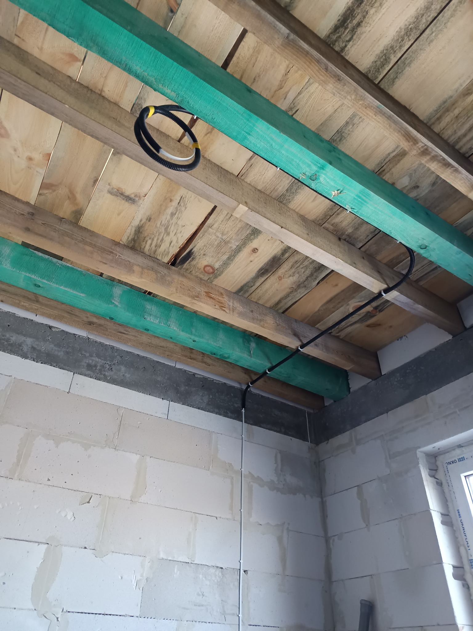 Czarny peszel z kablami przymocowany do drewnianej konstrukcji stropu w surowym wnętrzu budynku z oknem. Widoczne białe ściany z betonu komórkowego.