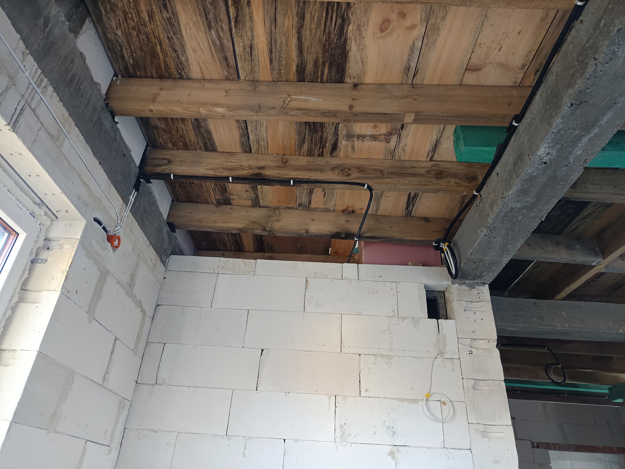 Wnętrze w trakcie budowy z widoczną instalacją elektryczną poprowadzoną po drewnianym stropie i ścianach z białych bloczków. Widoczne kable w peszlach i puszki instalacyjne.