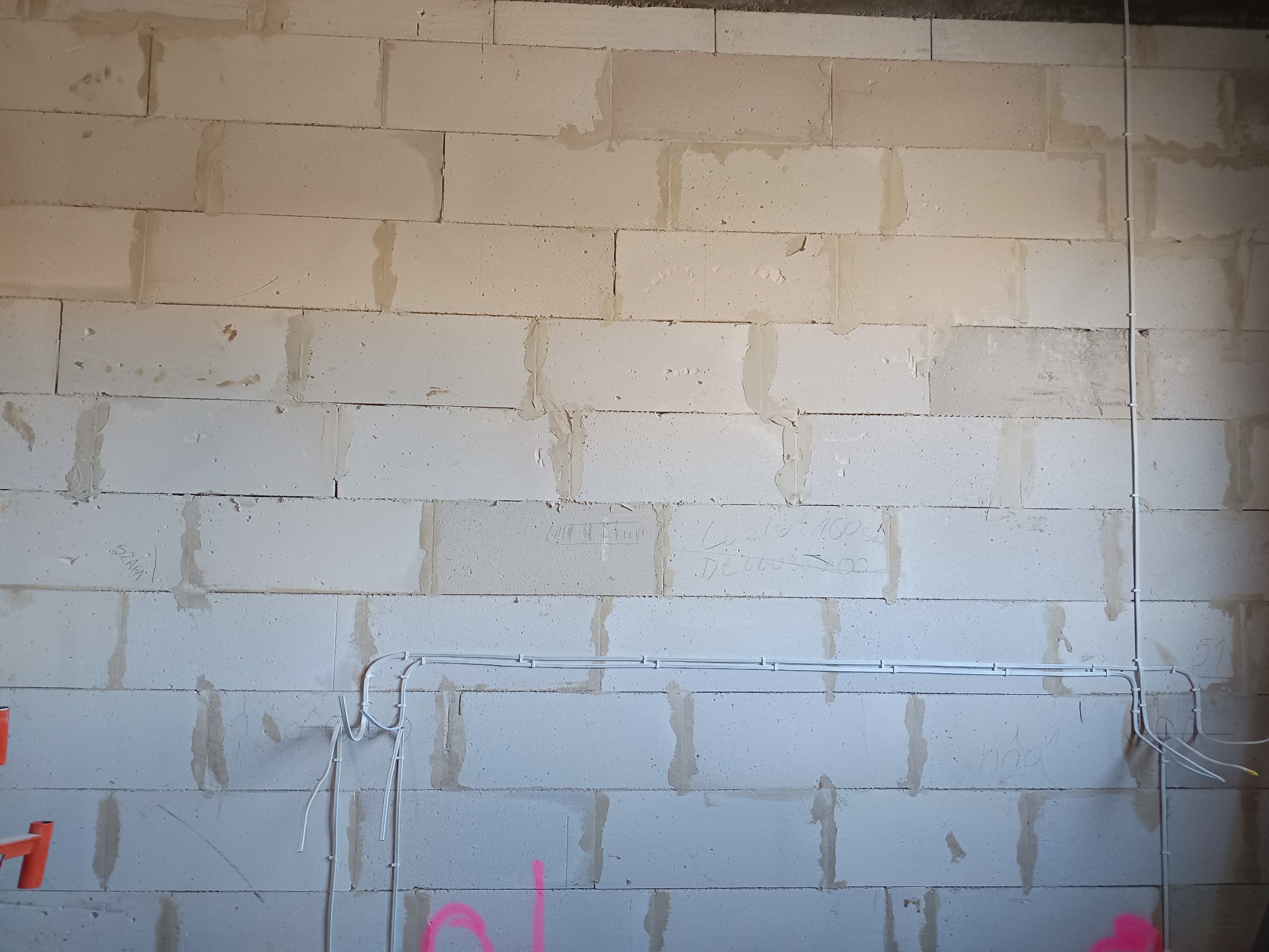 Biała ściana z betonu komórkowego z widocznymi, poprowadzonymi przewodami elektrycznymi w białych listwach. Widoczne napisy na ścianie.