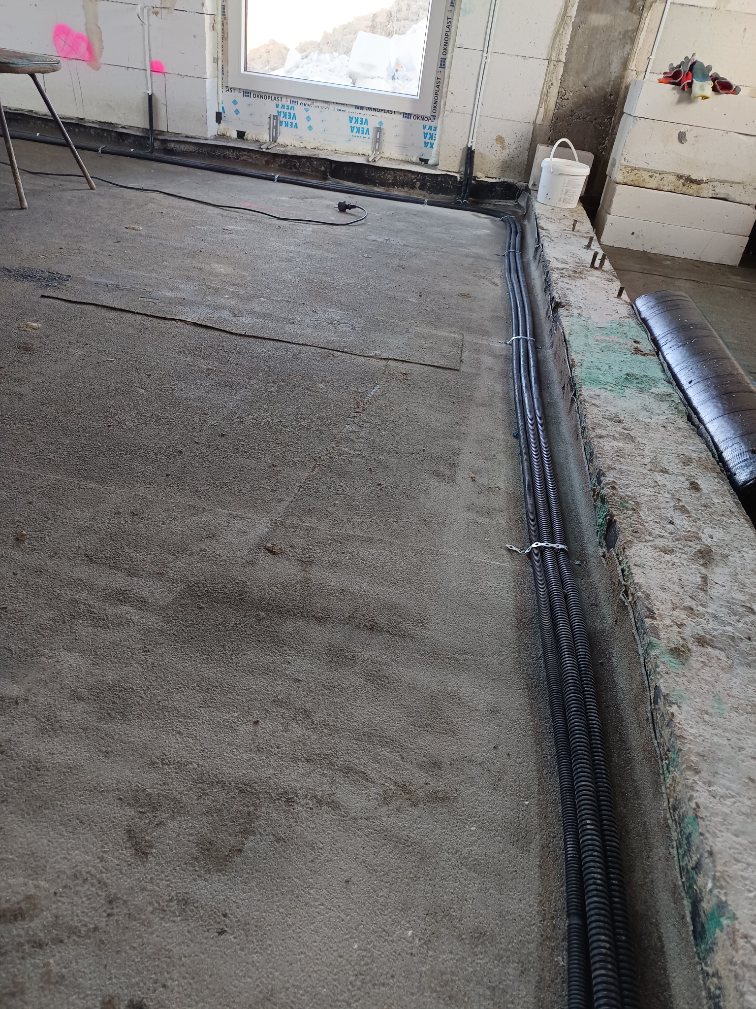 Kable w peszlach ułożone w bruzdach w podłodze, przygotowane do zalania wylewką. Widoczne narzędzia i materiały budowlane. Etap prac instalacyjnych w surowym wnętrzu.