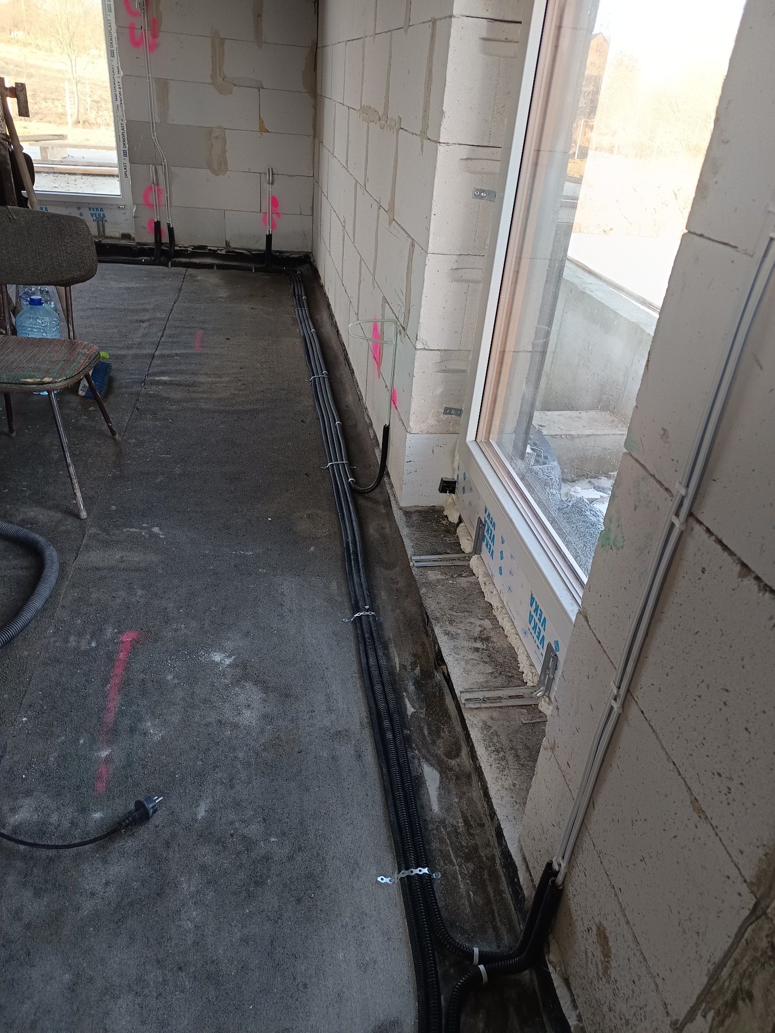 Wnętrze w trakcie prac instalacyjnych: kable elektryczne w peszlach ułożone wzdłuż ściany z betonu komórkowego, okno z widoczną izolacją. Oznaczenia na ścianach.