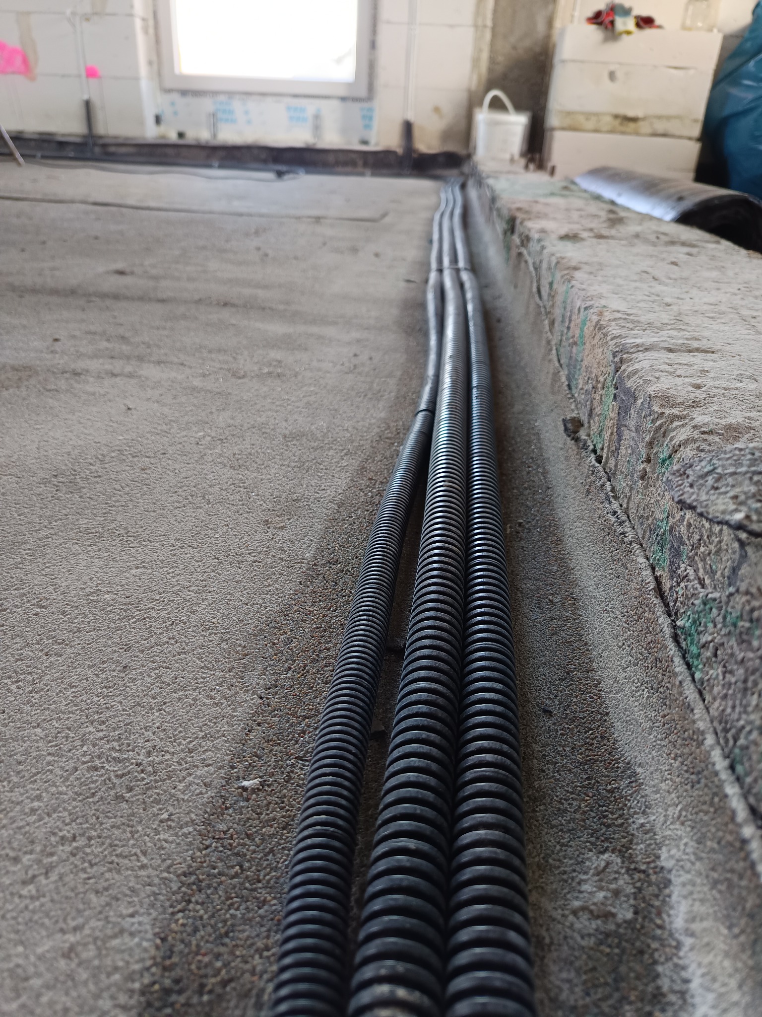 Trzy czarne rury karbowane ułożone równolegle na szarej, betonowej podłodze w surowym wnętrzu budynku w trakcie prac instalacyjnych. Widoczne okno w tle.
