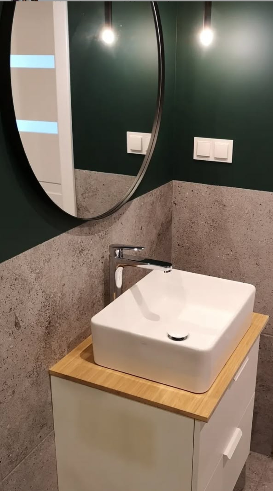 Nowoczesna łazienka z umywalką nablatową na szafce z drewnianym blatem, okrągłe lustro na ciemnozielonej ścianie, szara ściana wyłożona płytkami imitującymi beton.