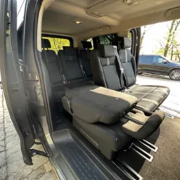 Wnętrze przestronnego busa z rozłożonymi fotelami, widok z otwartych drzwi na ulicę i zaparkowany samochód.