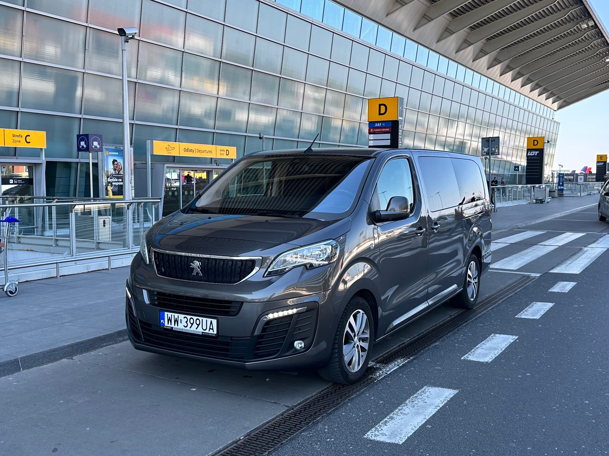 Szary bus Peugeot Traveller z polską rejestracją WW 399UA zaparkowany przed terminalem lotniska w słoneczny dzień, widoczne oznakowanie strefy D odlotów.