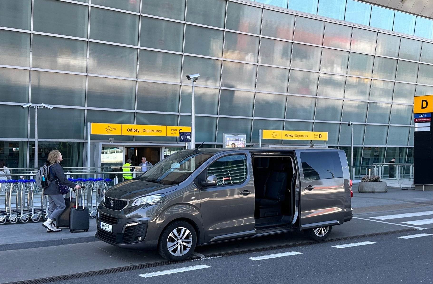 Szary bus Peugeot z otwartymi drzwiami stoi przed terminalem lotniska, widoczny znak 'Odloty/Departures', pasażerka z walizką w tle.