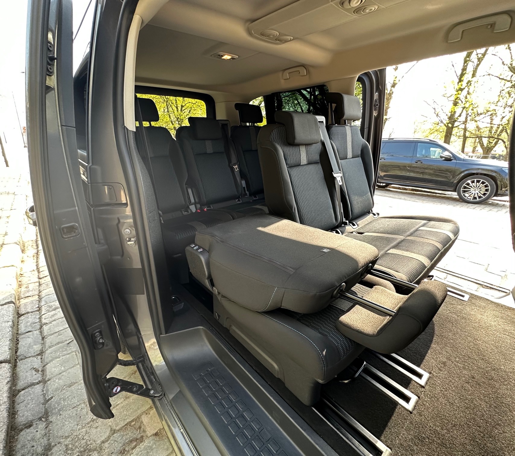 Wnętrze przestronnego busa z rozłożonymi fotelami, widok z otwartych drzwi na ulicę i zaparkowany samochód.