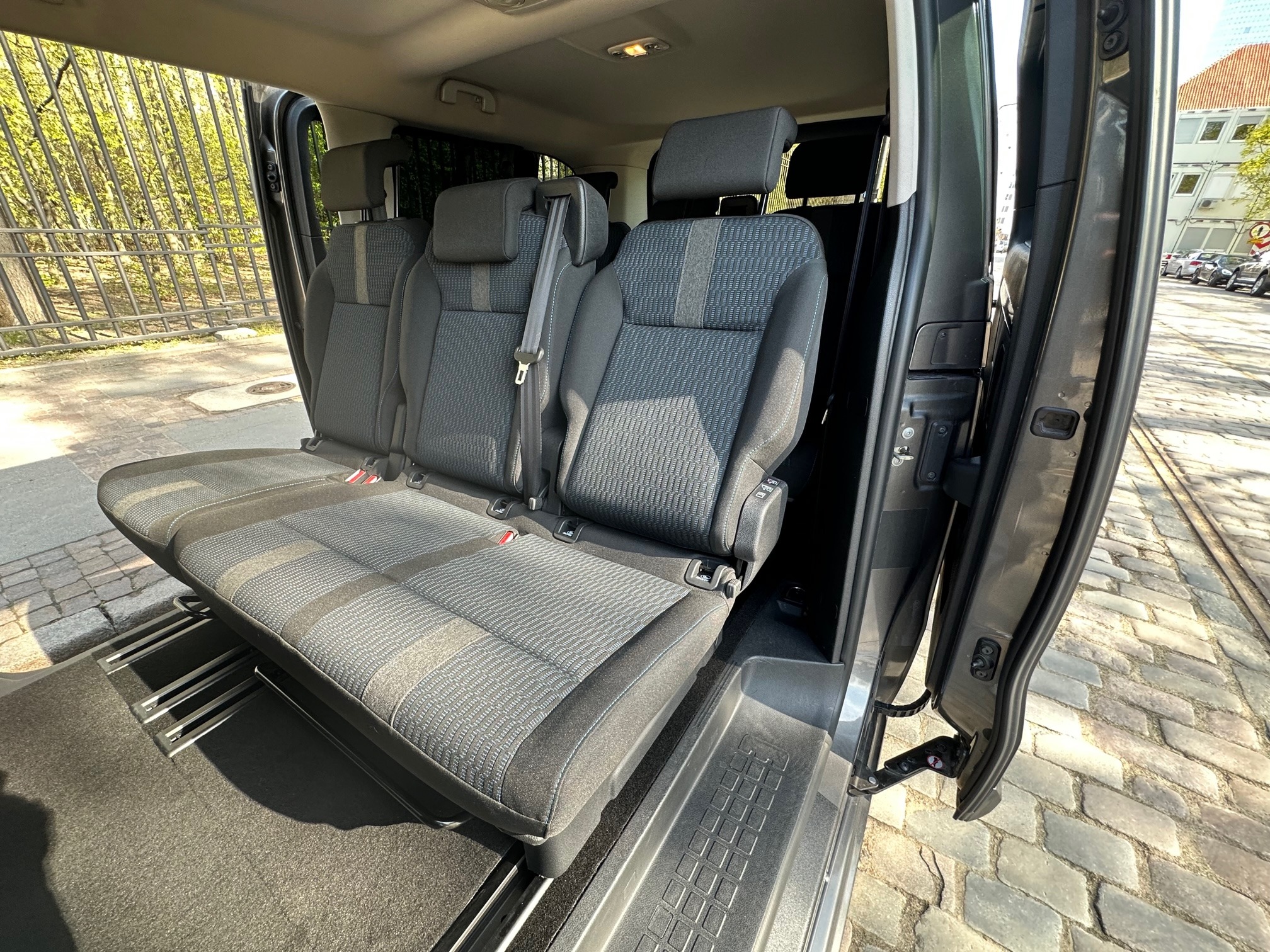 Wnętrze szarego busa z trzema fotelami z tapicerką w geometryczny wzór, widok z otwartych drzwi na ulicę z kostki brukowej i budynek w tle.