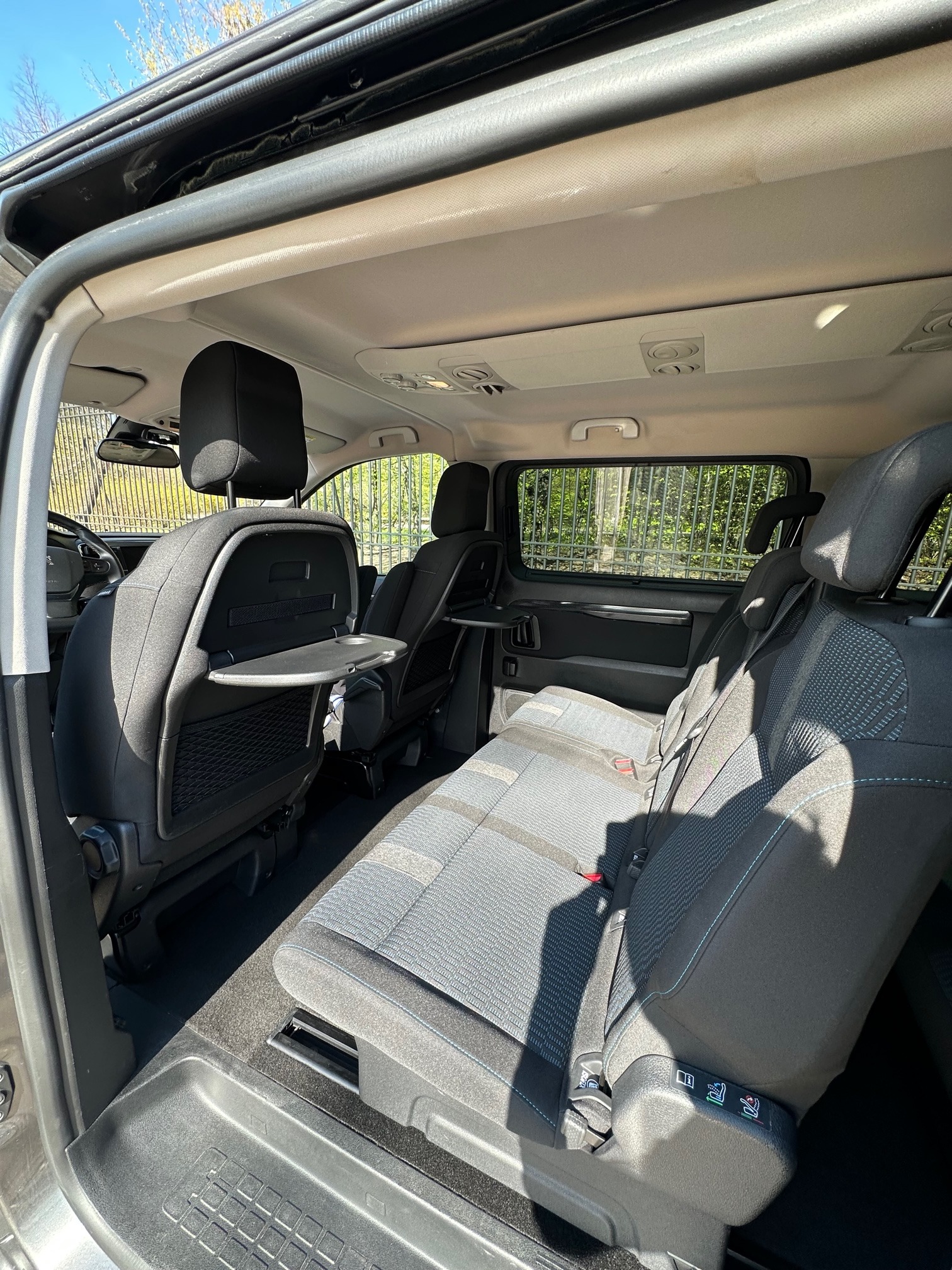 Wnętrze przestronnego busa z rozkładanymi stolikami przy tylnych siedzeniach, widok z otwartych drzwi, jasna tapicerka z niebieskimi akcentami, w tle zieleń drzew za oknem.