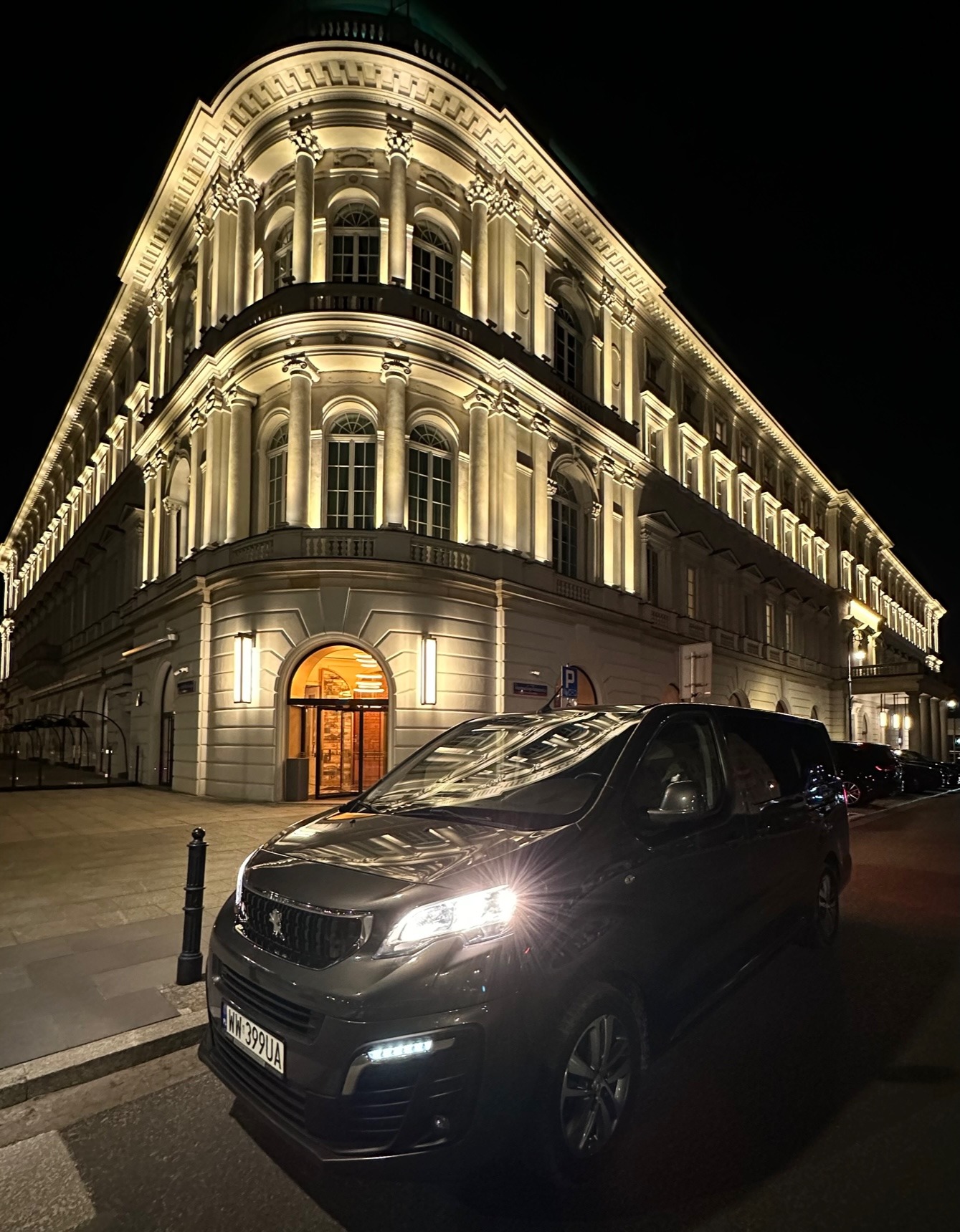 Czarny bus Peugeot Expert z włączonymi światłami, zaparkowany nocą na tle oświetlonego klasycystycznego budynku w Warszawie.