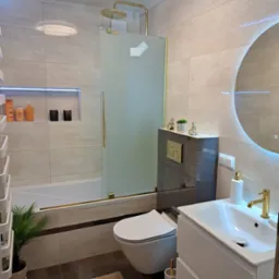 Nowoczesna łazienka po remoncie w Lublińcu: beżowe płytki, szklany parawan nawannowy ze złotymi detalami, okrągłe lustro z podświetleniem LED.