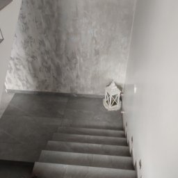 Glazurowy Kwadrat - Schody wyłożone szarymi płytkami, ściana z dekoracyjną szarą strukturą, biały lampion dekoracyjny. Widok z góry na dół, stonowana kolorystyka.