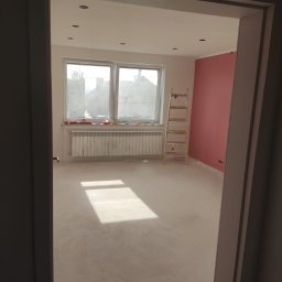Glazurowy Kwadrat - Pokój po szpachlowaniu, przygotowany do malowania. Widoczne gładkie ściany, okno z grzejnikiem i drabina malarska. Świeżo wykończone wnętrze z różową ścianą.