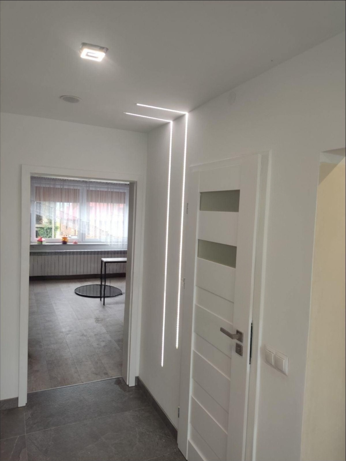 Białe wnętrze: drzwi z przeszkleniami, podłoga z szarych płytek, designerskie oświetlenie LED wzdłuż ściany. Widok na pokój z oknem i grzejnikiem.