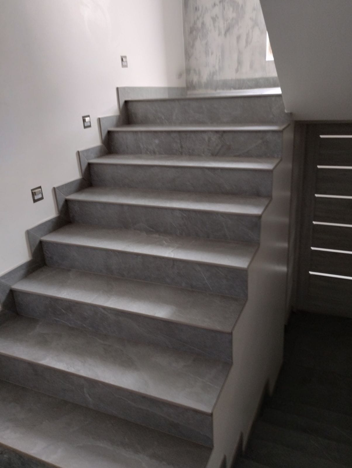 Schody wyłożone szarymi płytkami imitującymi marmur, z subtelnym oświetleniem punktowym na ścianie. Widoczny fragment drzwi w tle. Ujęcie z dołu, podkreślające geometrię.