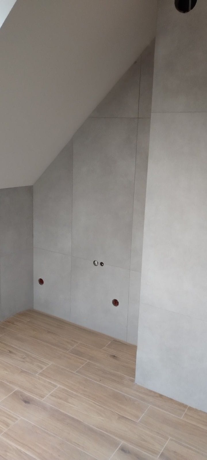 Wykończona łazienka na poddaszu: szare płytki ścienne, drewnopodobne płytki podłogowe, widoczne otwory na gniazdka elektryczne i oświetlenie sufitowe.