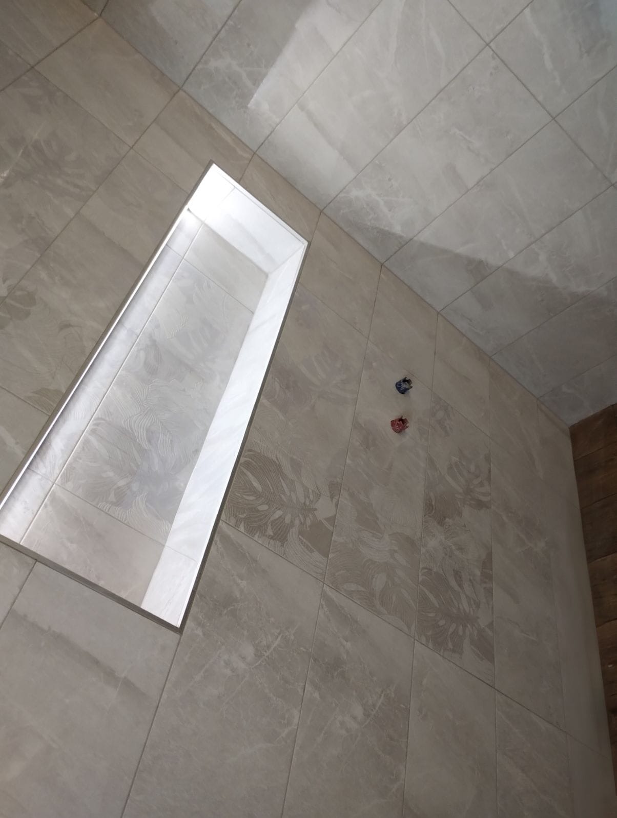 Wykończona łazienka z beżowymi płytkami imitującymi marmur, z wnęką prysznicową. Widoczne przyłącza wodne. Ukośne ujęcie podkreśla wzór płytek.