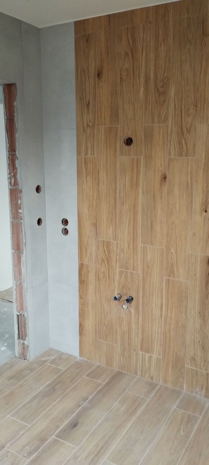 Łazienka w trakcie wykańczania: drewnopodobne płytki na ścianie i podłodze, widoczne otwory na instalacje. Częściowo wykończona ściana z szarych płytek.