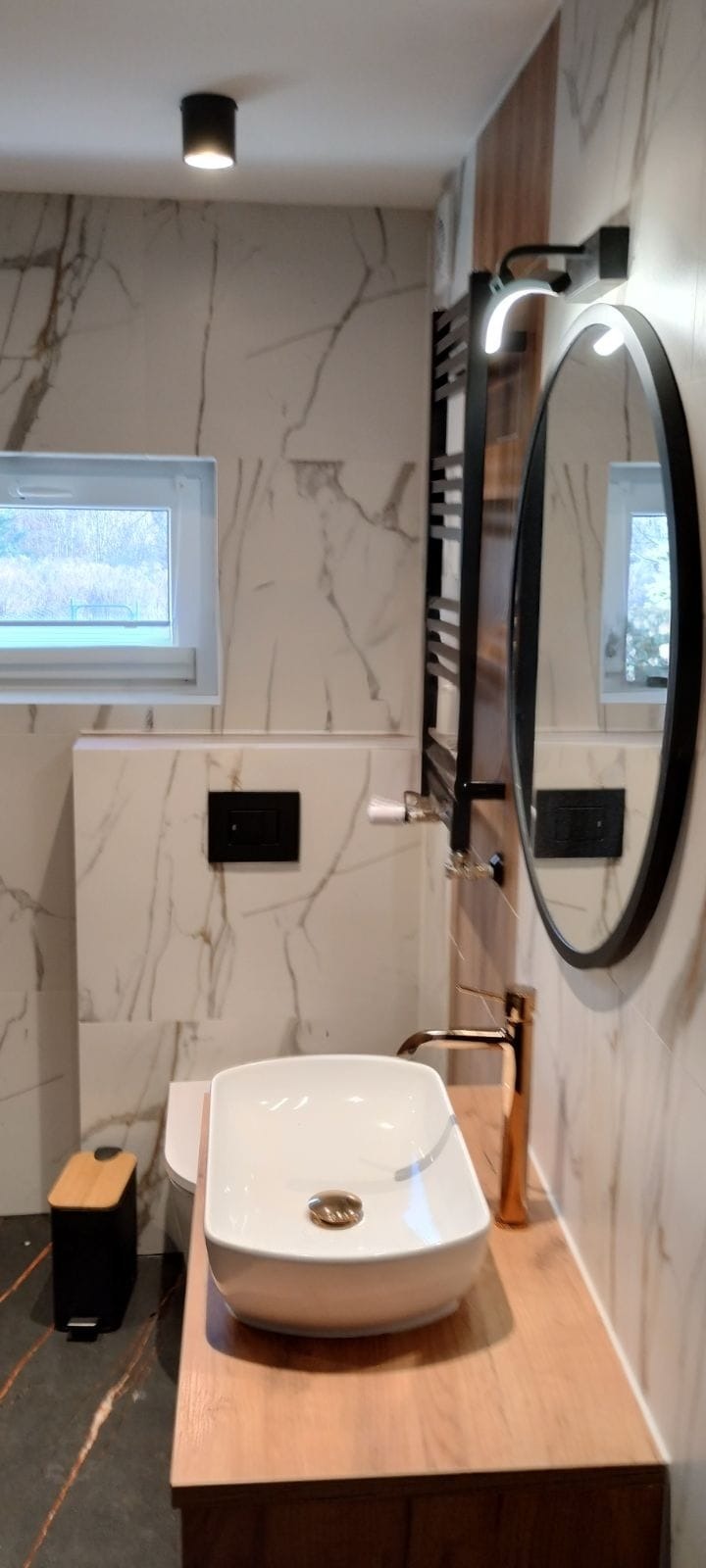 Nowoczesna łazienka z umywalką nablatową, baterią w kolorze różowego złota i marmurowymi płytkami. Widoczne okno i czarny grzejnik.