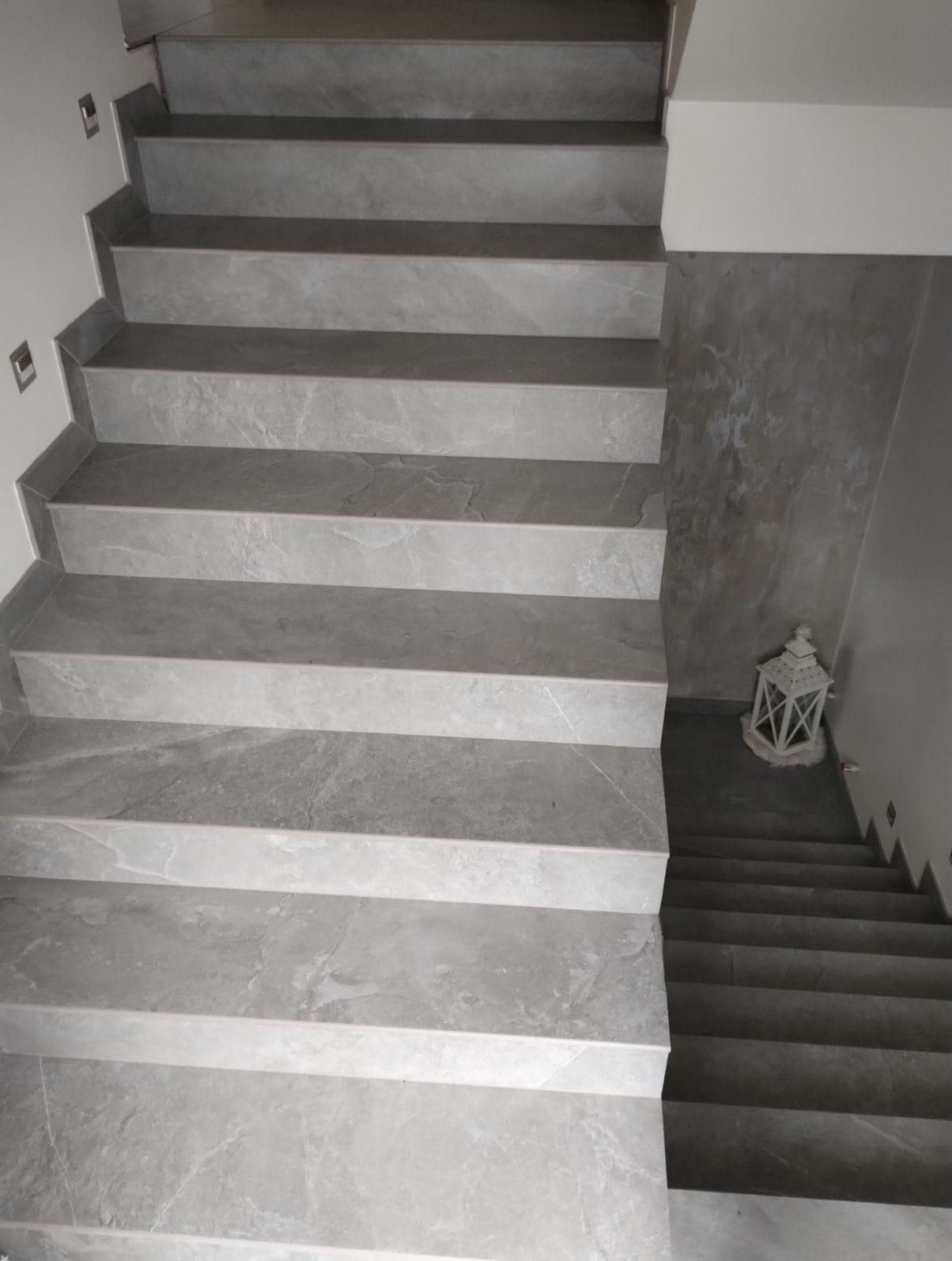 Wykończone schody z szarych płytek imitujących marmur, z dekoracyjną latarenką na podeście. Widok z góry, podkreślający wzór i precyzję ułożenia płytek.