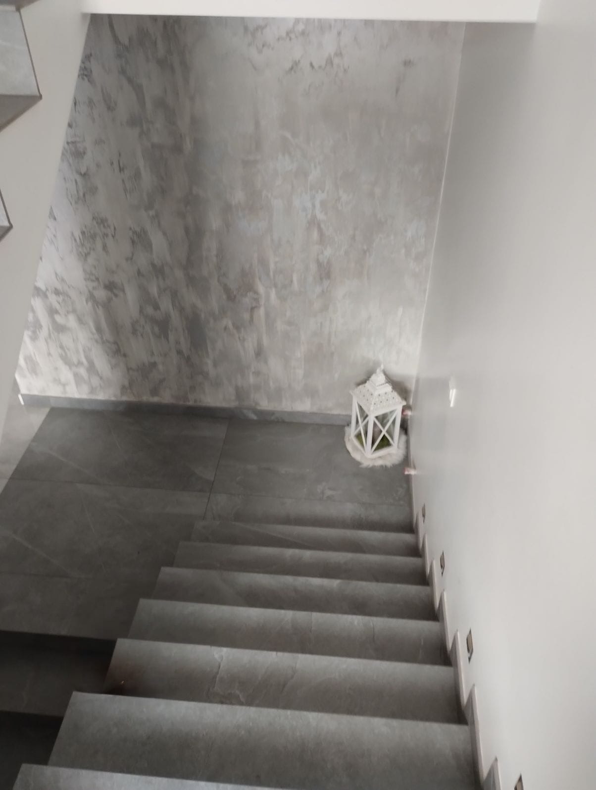 Schody wyłożone szarymi płytkami, ściana z dekoracyjną szarą strukturą, biały lampion dekoracyjny. Widok z góry na dół, stonowana kolorystyka.