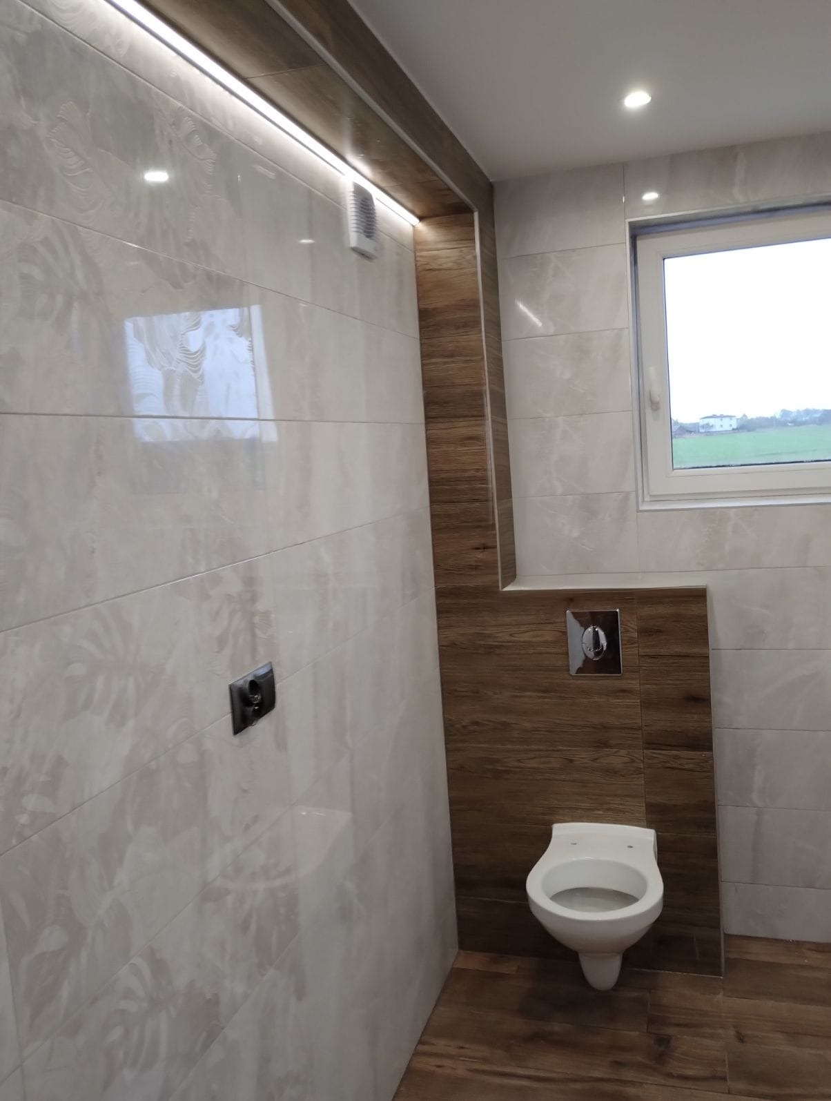 Nowoczesna łazienka z beżowymi płytkami imitującymi marmur, drewnopodobnymi akcentami i podświetleniem LED. Widoczny podwieszany sedes i okno z widokiem na zieleń.