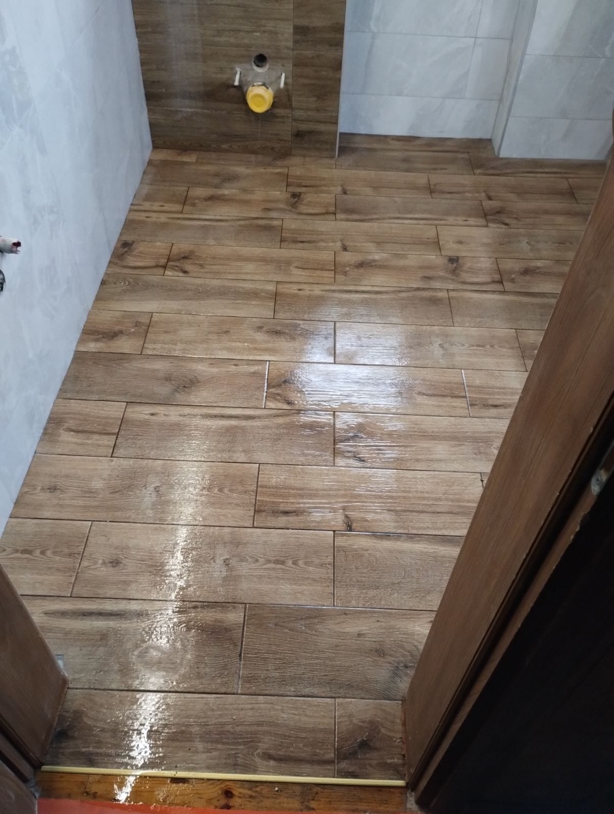 Ukończona podłoga w łazience wyłożona brązowymi płytkami imitującymi drewno, widok z góry, z fragmentem drzwi po prawej stronie i instalacją sanitarną na ścianie.